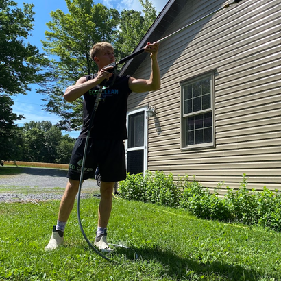 Person standing on grass, aiming a long tool upward beside a house on a sunny day