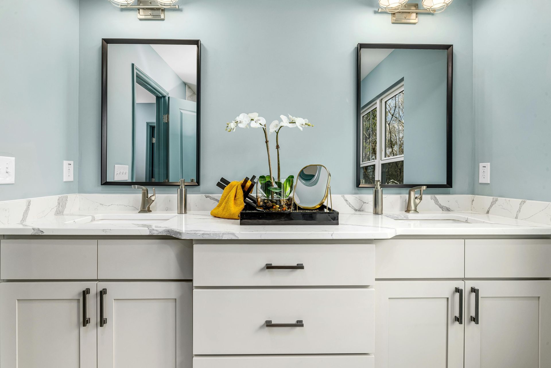 Bathroom with two sinks, mirrors, white cabinets, and light blue walls.
