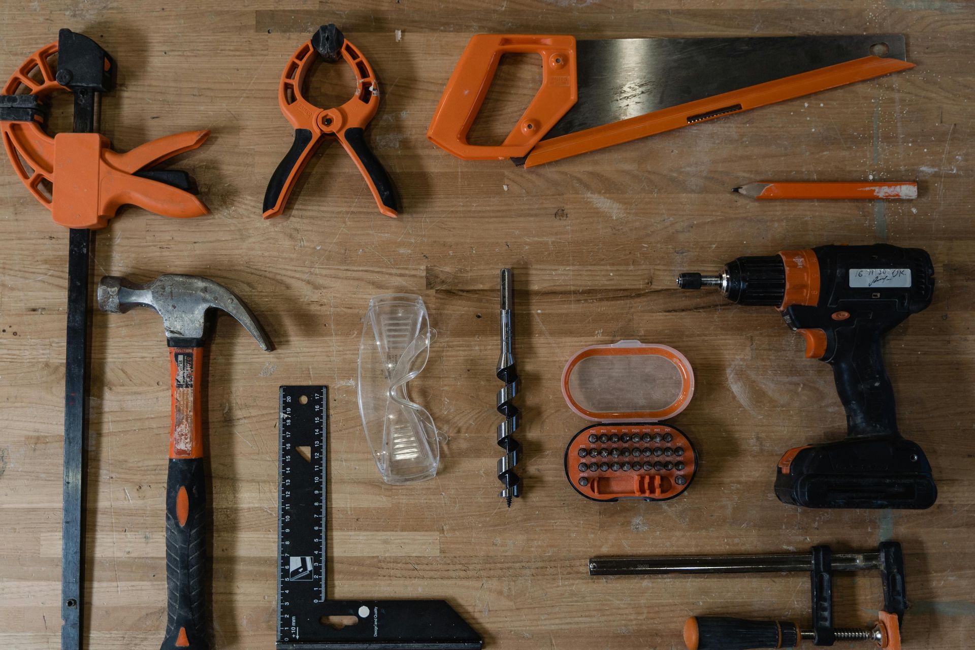 Arrangement of woodworking tools: clamps, saw, pencil, drill, hammer, safety glasses, drill bits, square, on wooden surface.