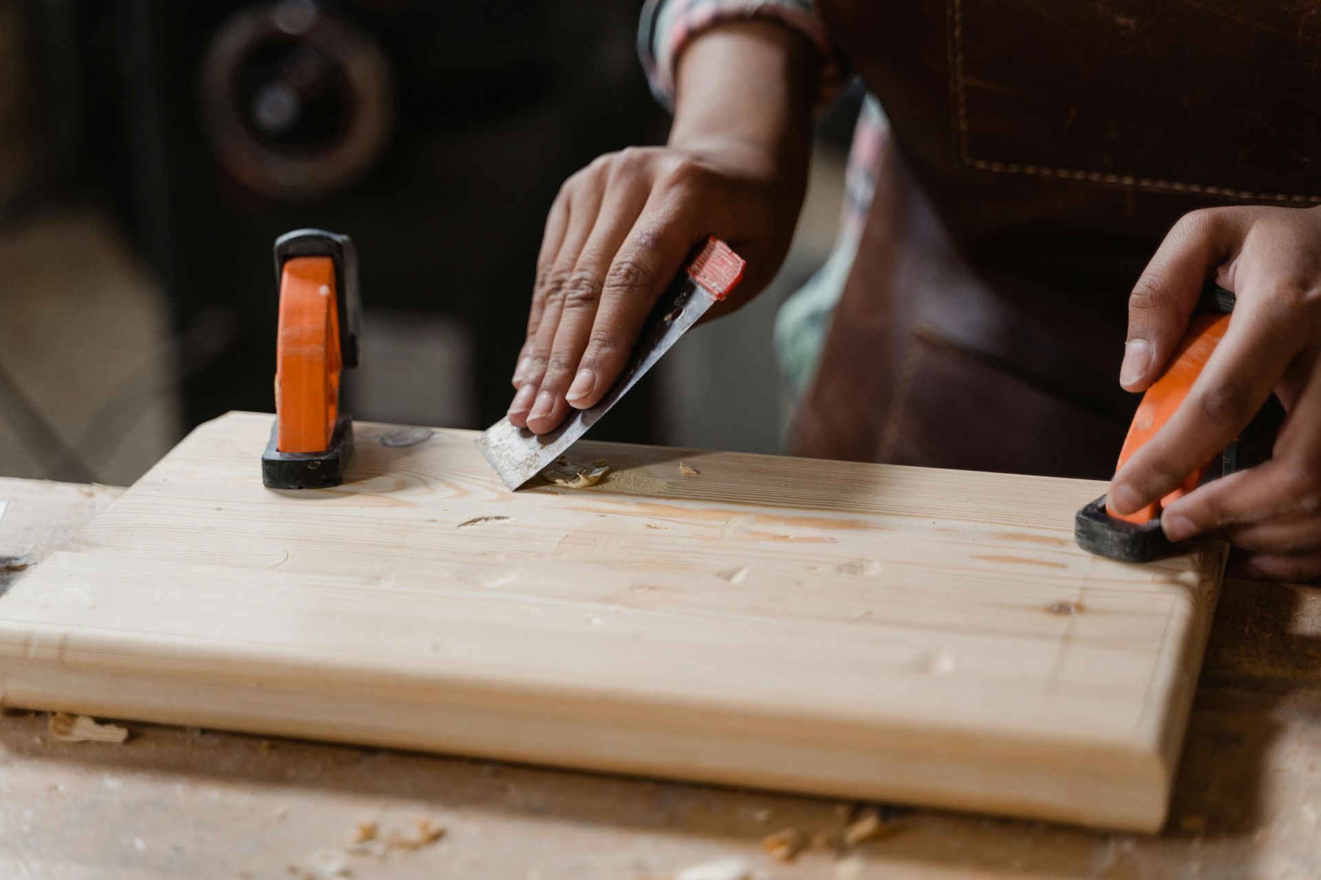 Person using a chisel to shape a wooden plank, held in place by clamps, in a workshop.