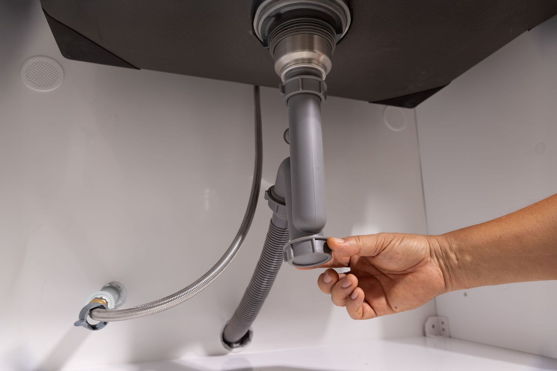 Person's hand unscrews a drain trap under a white sink, near a gray flexible drain pipe.