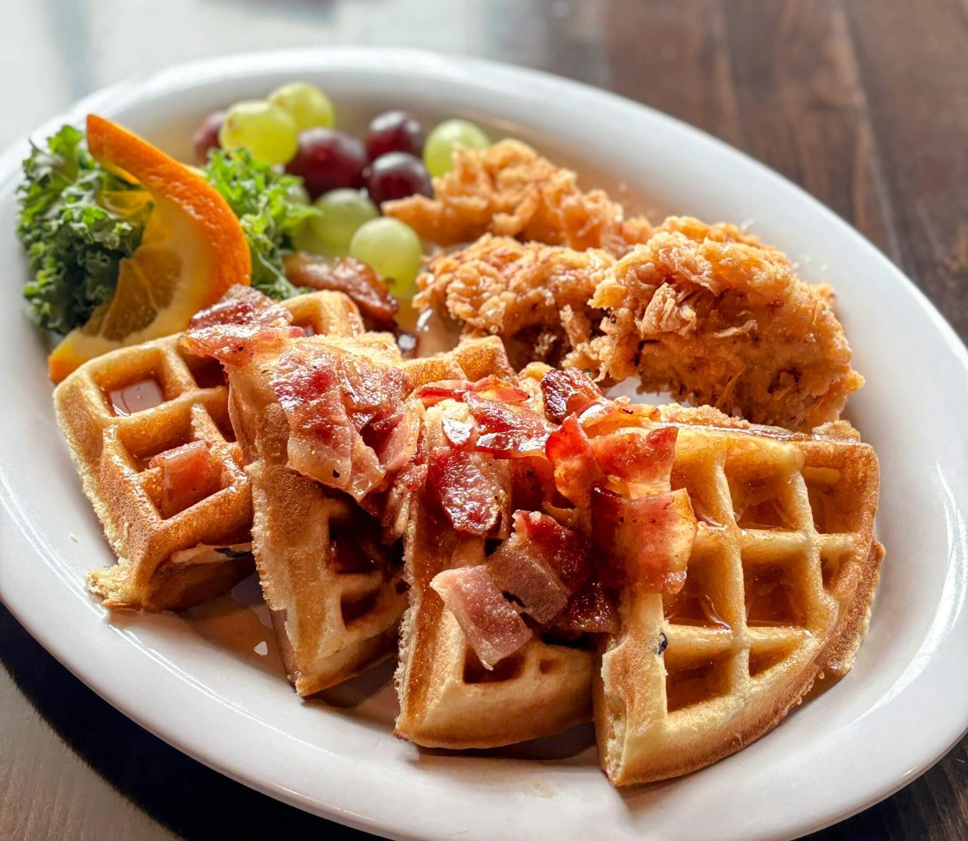 A white plate topped with waffles , bacon , chicken and fruit.