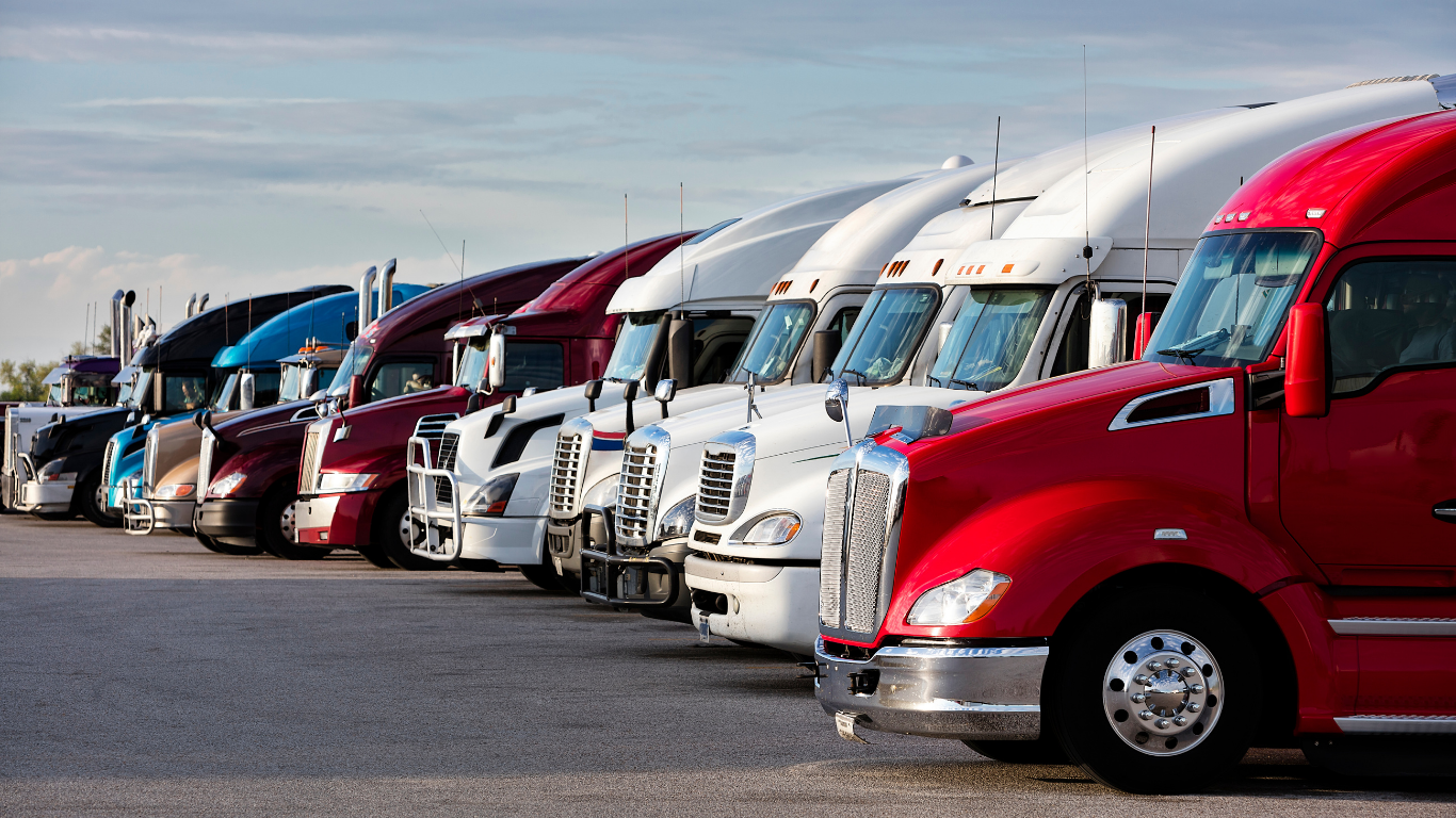 Semi-trucks of various colors parked in a row on a paved lot under a cloudy sky.