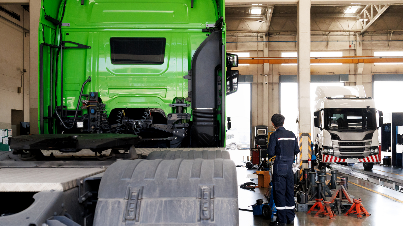 A mechanic inspects a green semi-truck in a repair shop, another truck in the background.