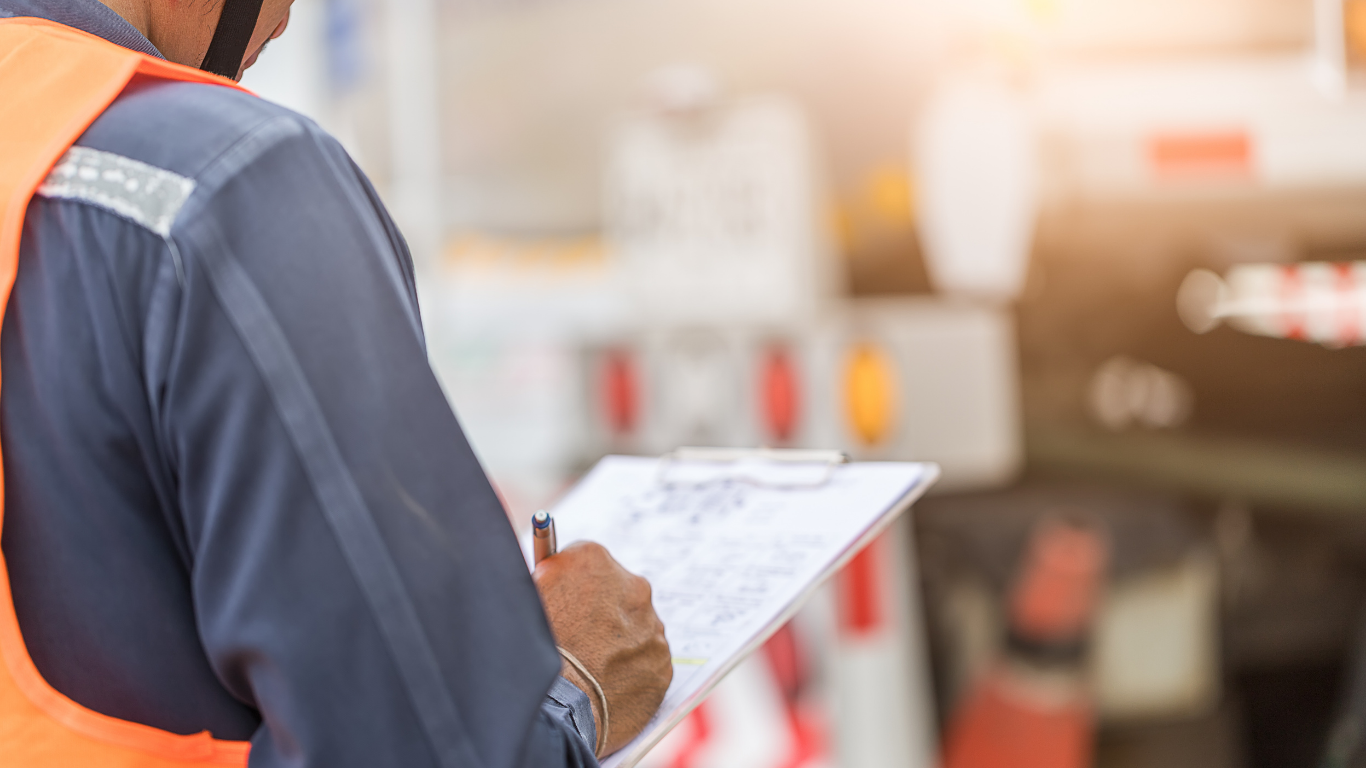 Person in safety vest writes on clipboard near road construction equipment.