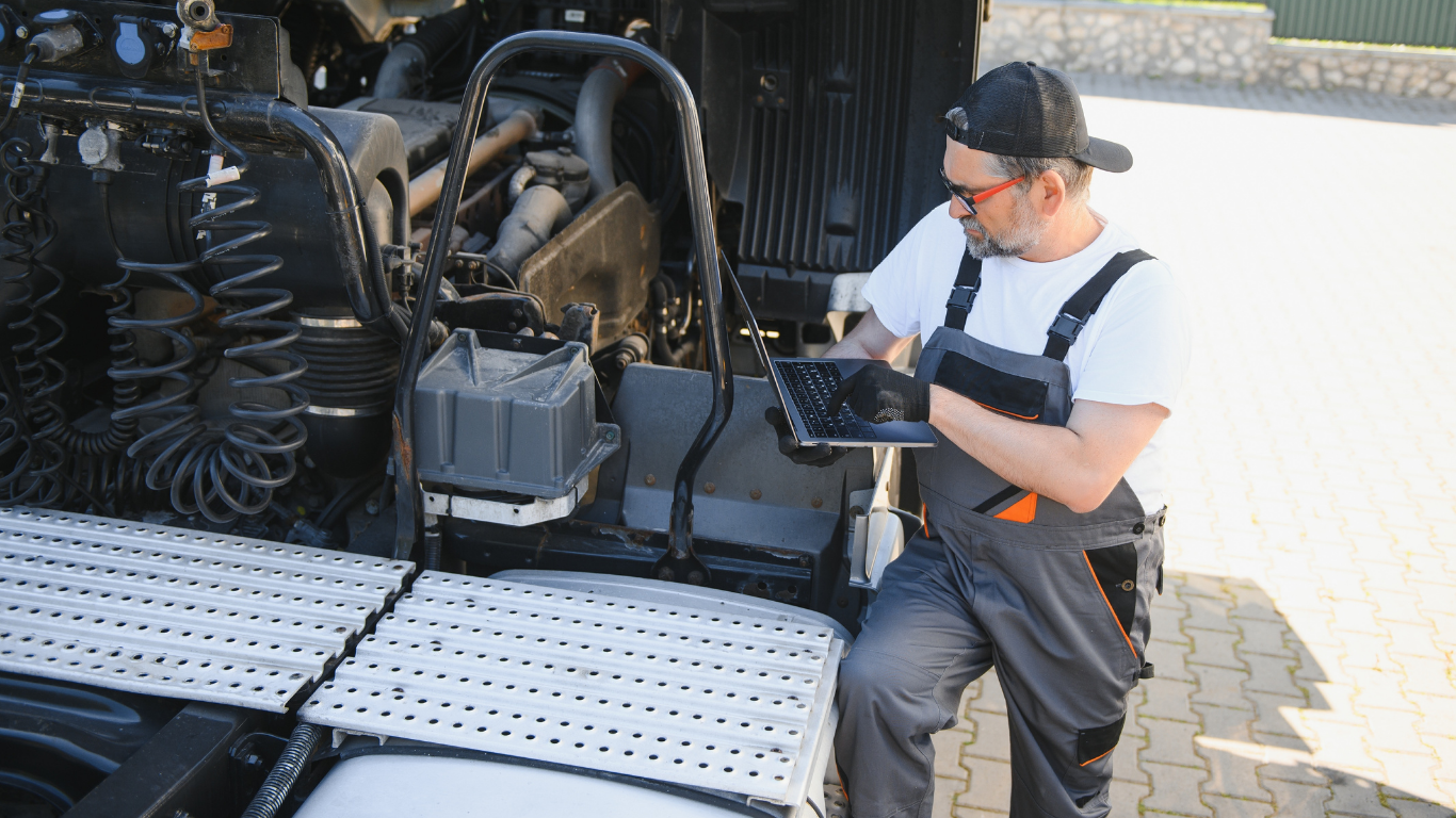 Mechanic in overalls uses a tablet to inspect a truck engine. Sunny outdoor setting.