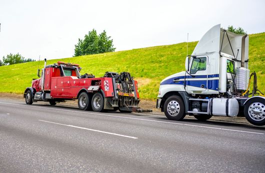 Red tow truck towing a white and blue semi-truck on a road, with green hill in the background.