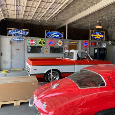 Classic cars in a garage with vintage signs, including Chevy and Ford, and a red pickup truck.