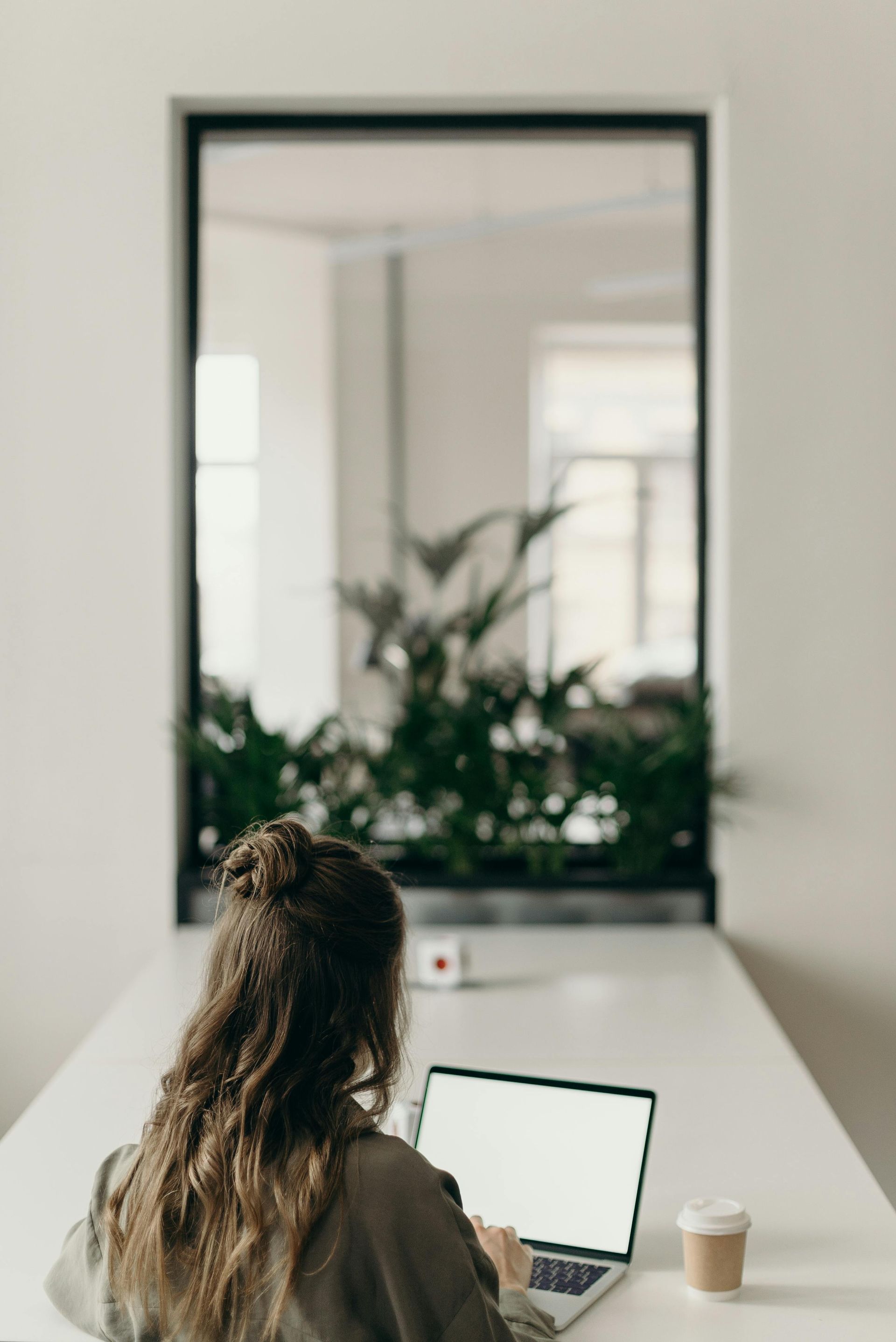 Woman at a white table, working on a laptop with a blank screen. A window with plants is in the background.