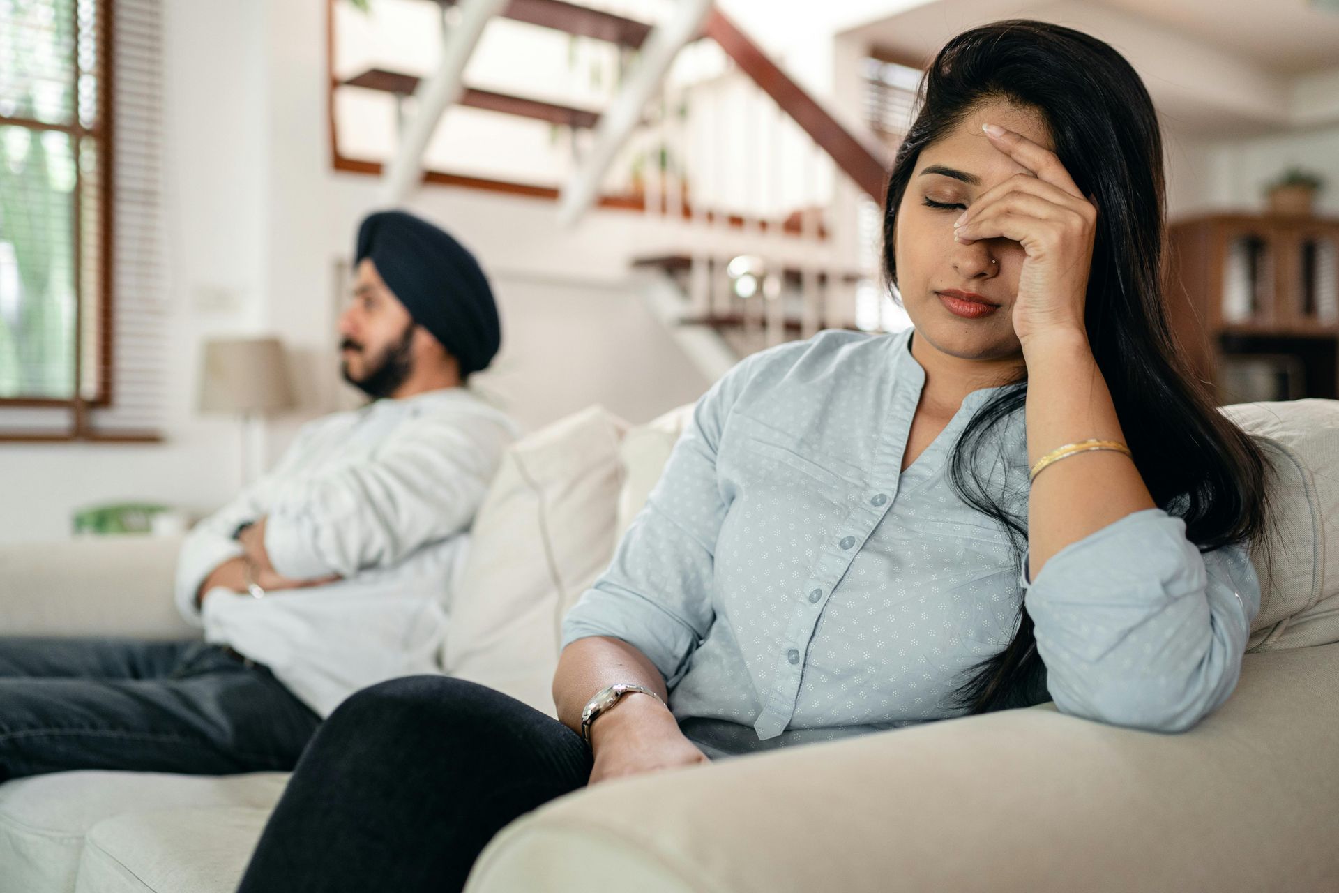 Woman with hand on forehead looking distressed, man in background, apart, on a couch.