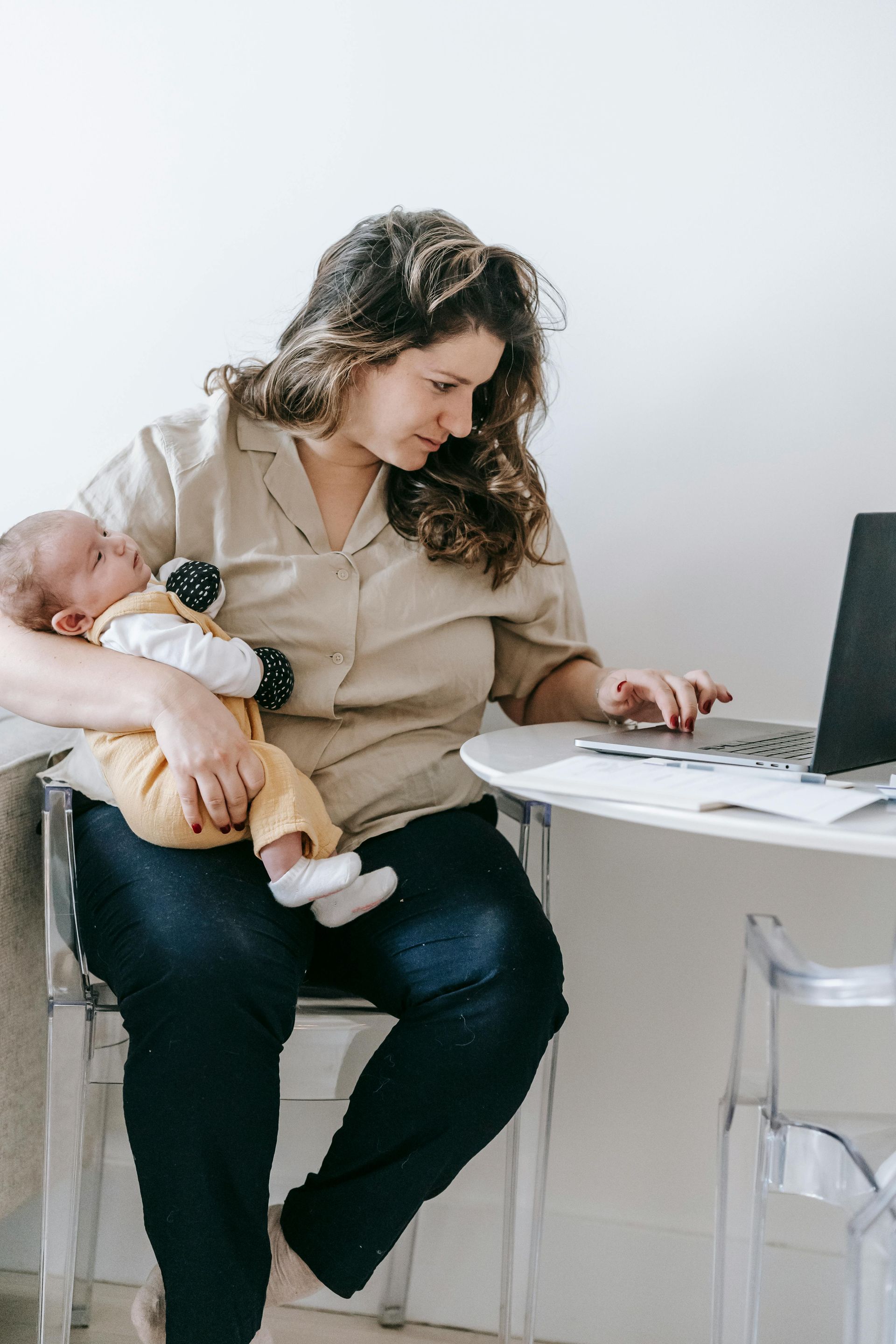 Woman with baby in arms working on laptop at a small white table.