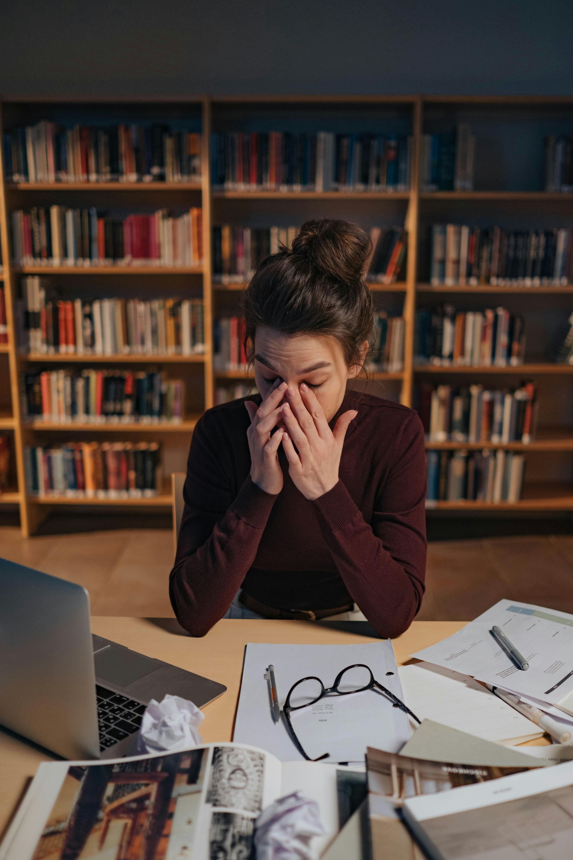 Woman stressed, studying at a library desk, laptop and papers scattered, rubbing eyes.