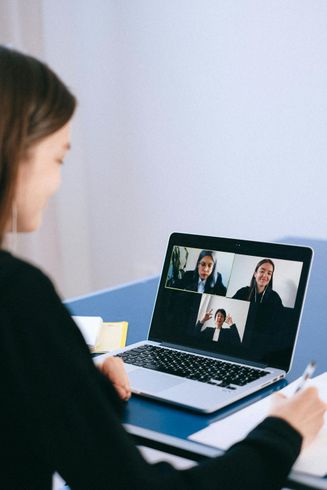 Woman on a video call, taking notes. Laptop shows three other participants, blue table, white wall.