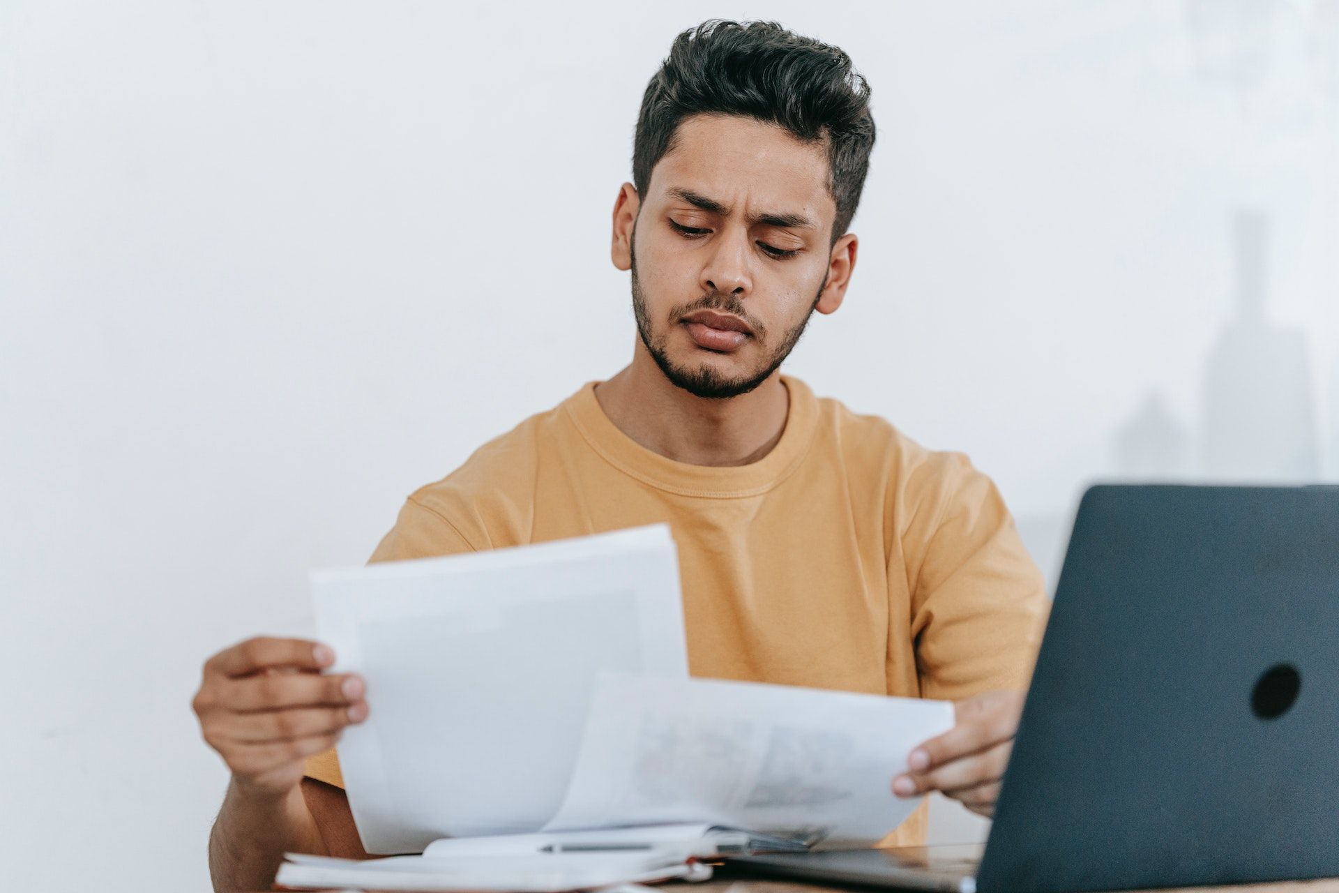 A person in a yellow shirt looks down at paper documents while sitting at a desk with a laptop.