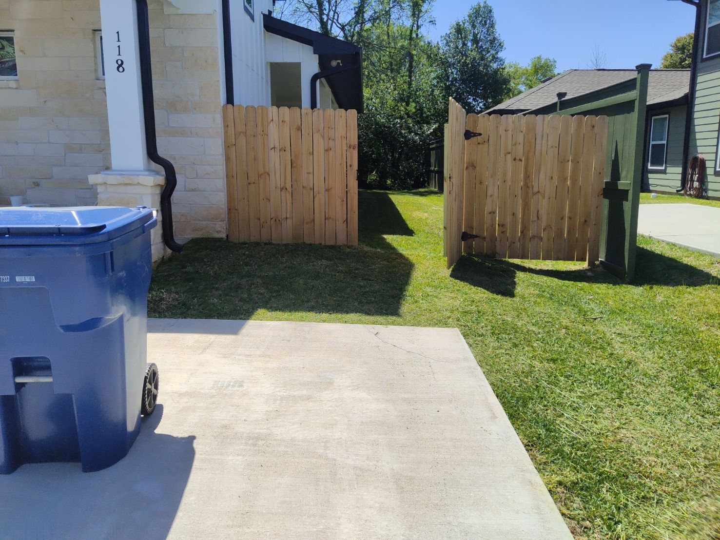 A blue trash can is parked in front of a house next to a wooden fence.