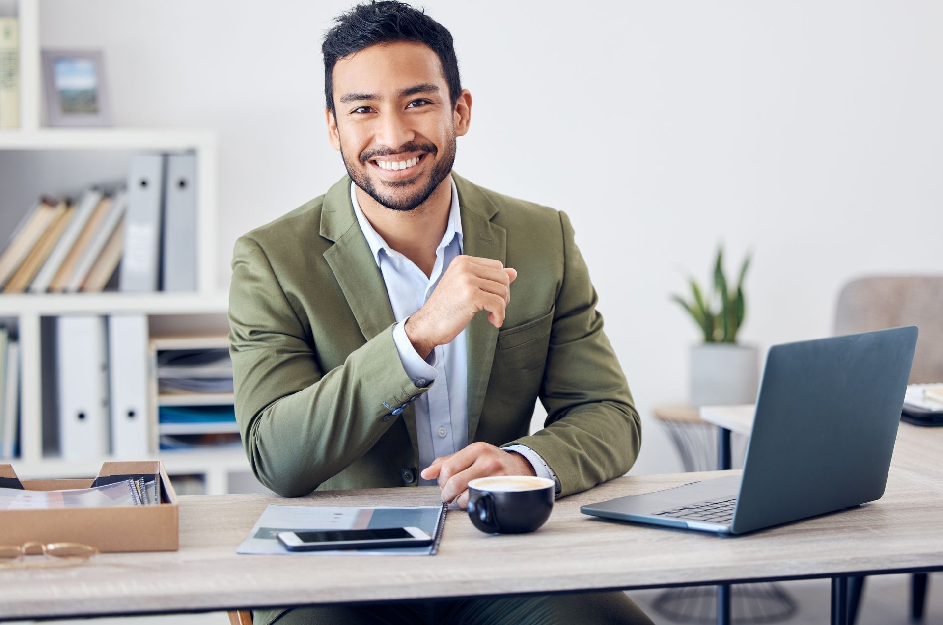 A man in a suit is sitting at a desk with a laptop and a cup of coffee.