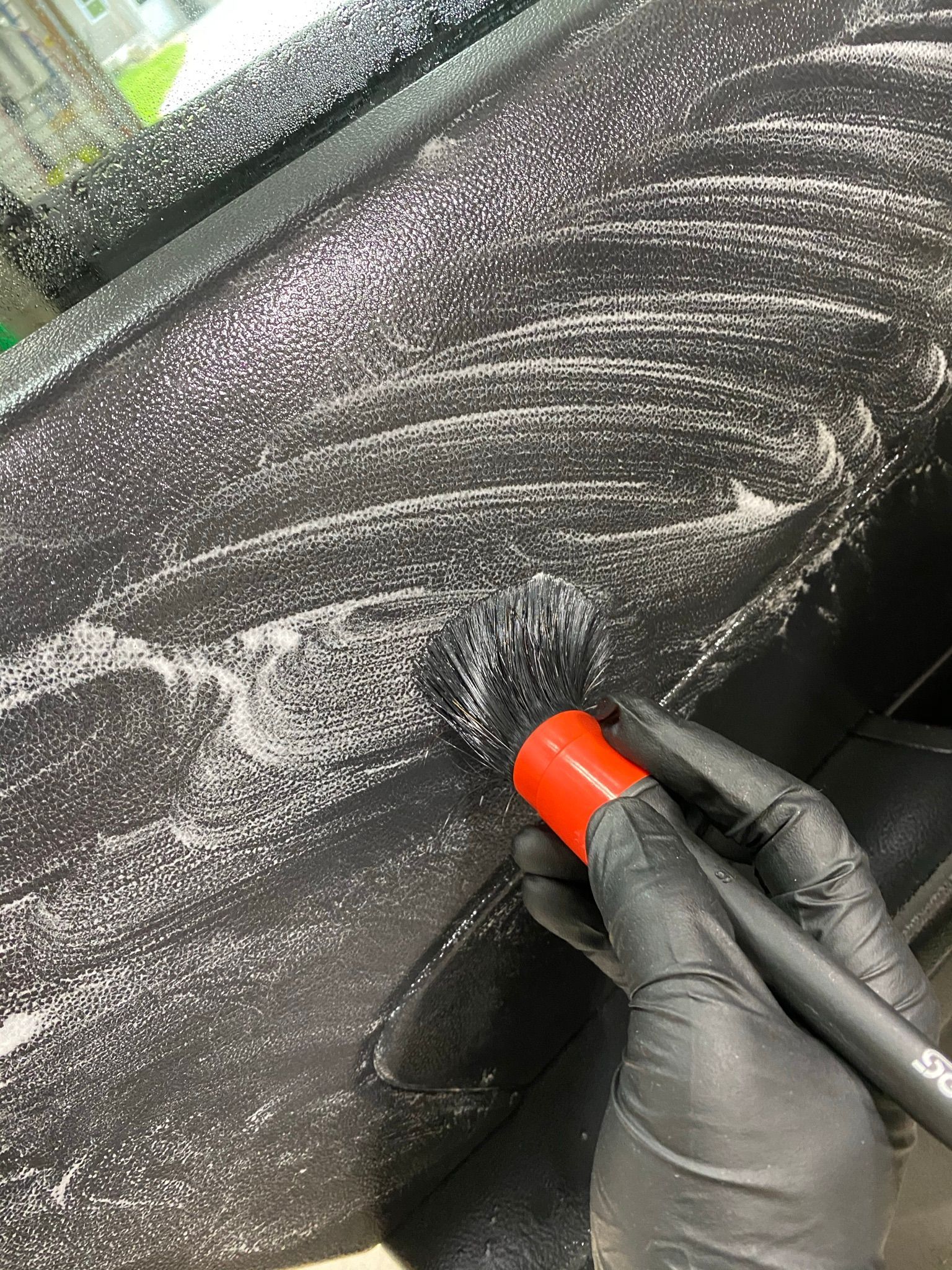Gloved hand using a brush to clean a car's interior, covered in soapy white foam.