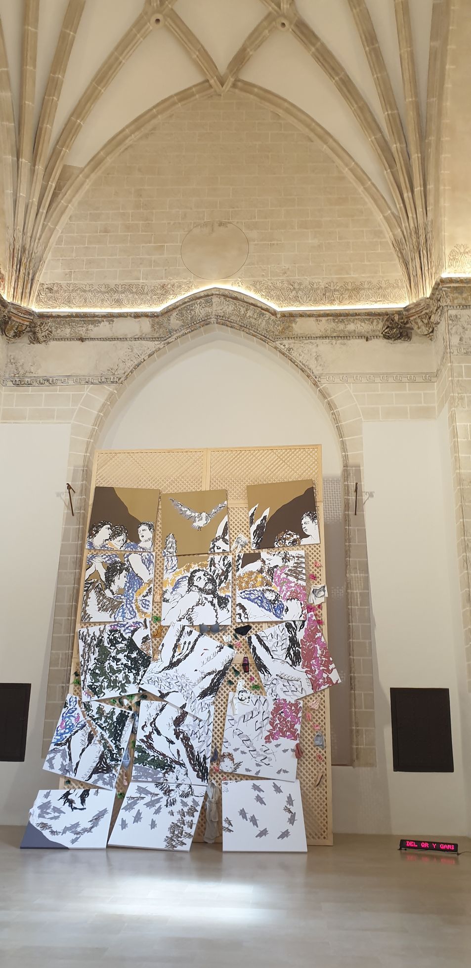 A large, fragmented collage of white paper with black ink sketches stands against a stone wall under vaulted ceilings.