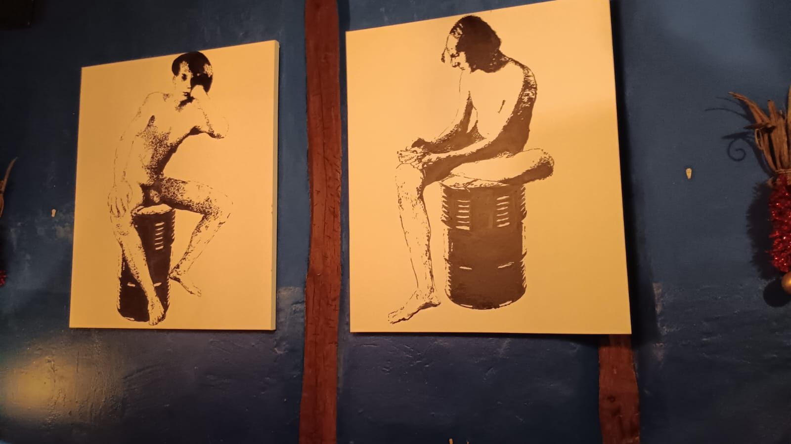 Two black and white paintings of seated figures on tan canvases against a blue wall.