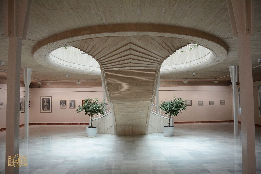 Grand gallery with a central staircase, two skylights, potted trees, and framed artwork on the walls.