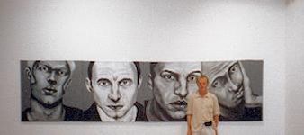 A man stands before a four-panel painting of gray-toned faces with varied expressions on a white wall.