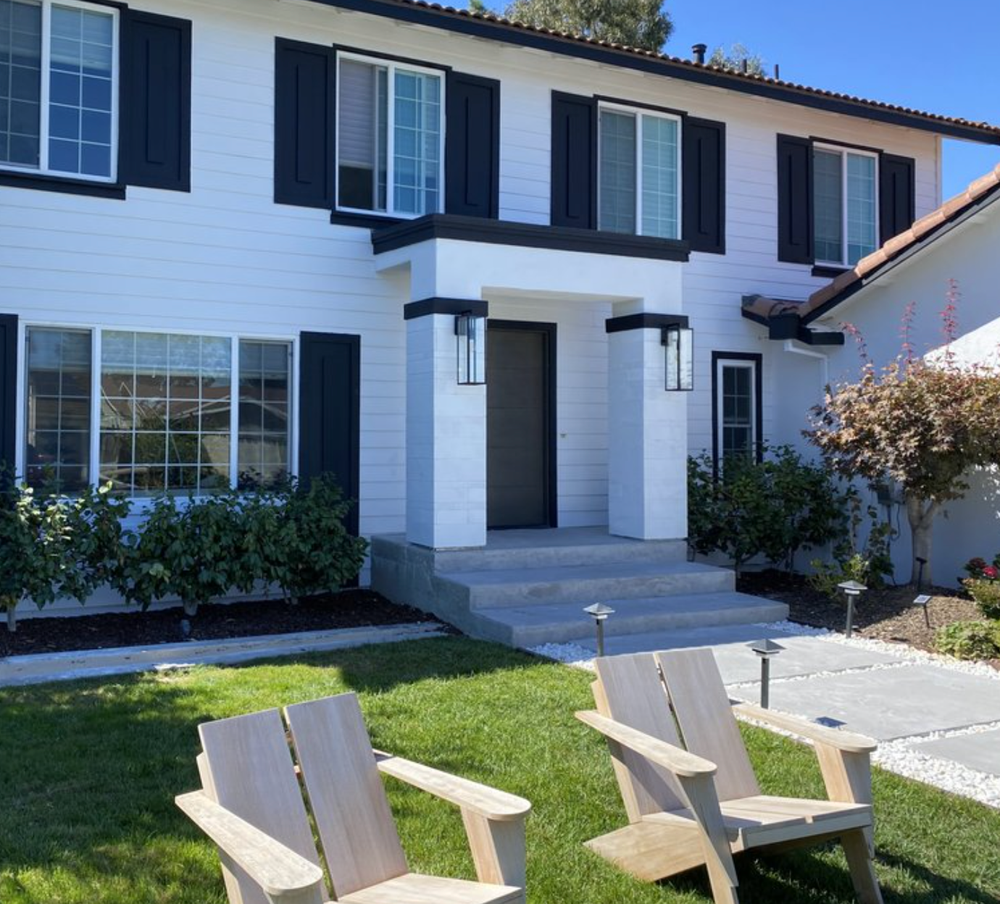 Two wooden chairs are sitting in front of a white house with black shutters.