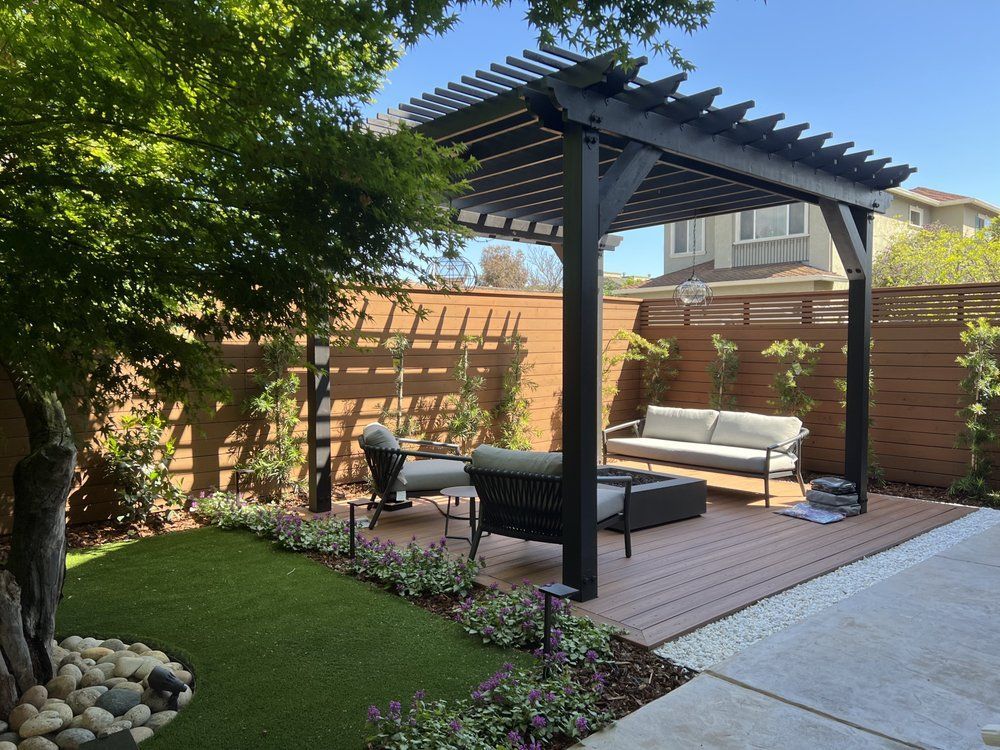 There is a pergola in the backyard with a couch and chairs underneath it.