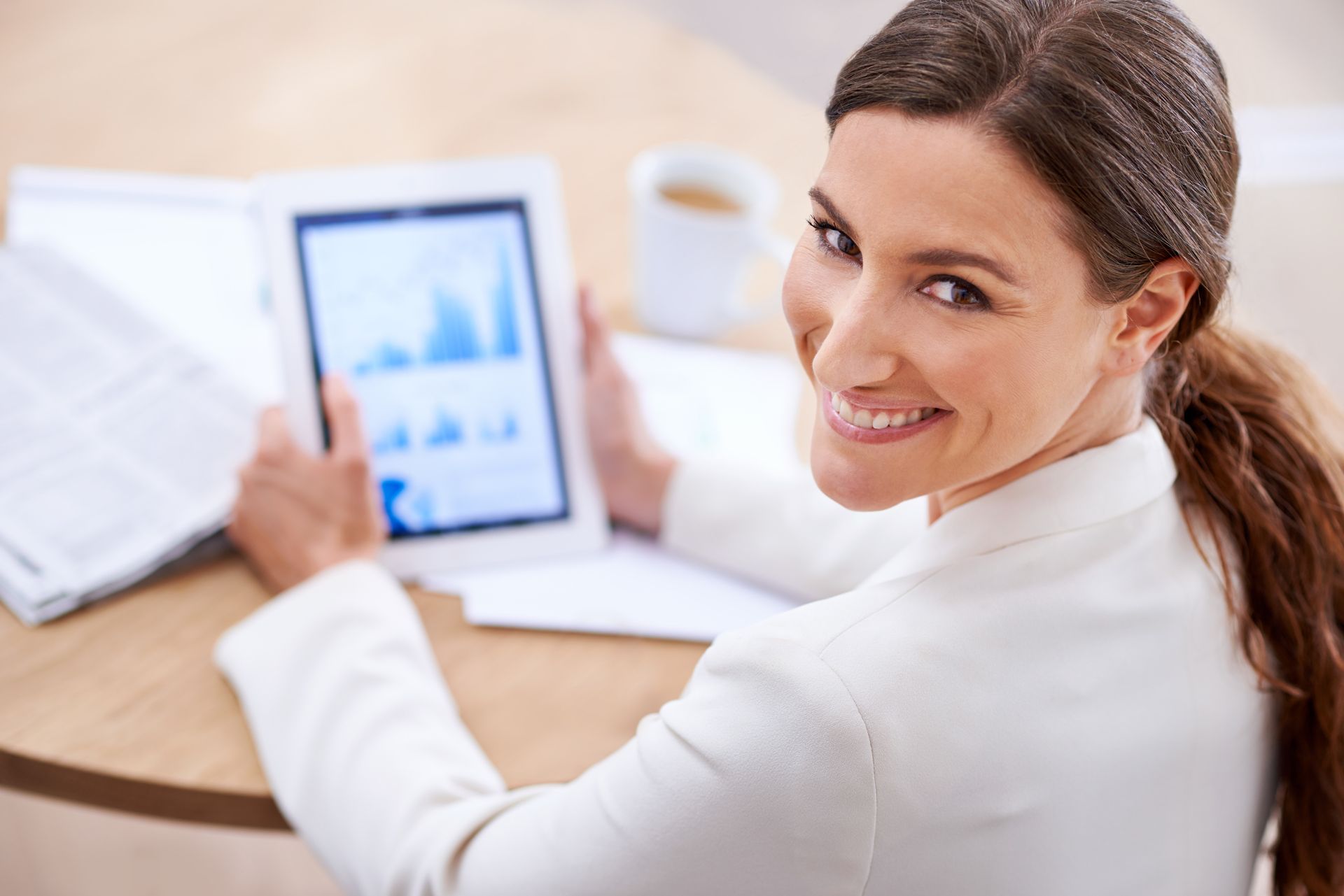 A woman is sitting at a table holding a tablet and smiling.