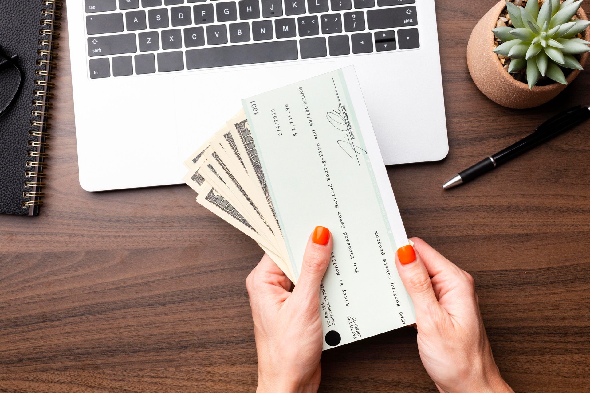 Hands holding a stack of checks on a wooden desk with a laptop, pen, notebook, and plant.