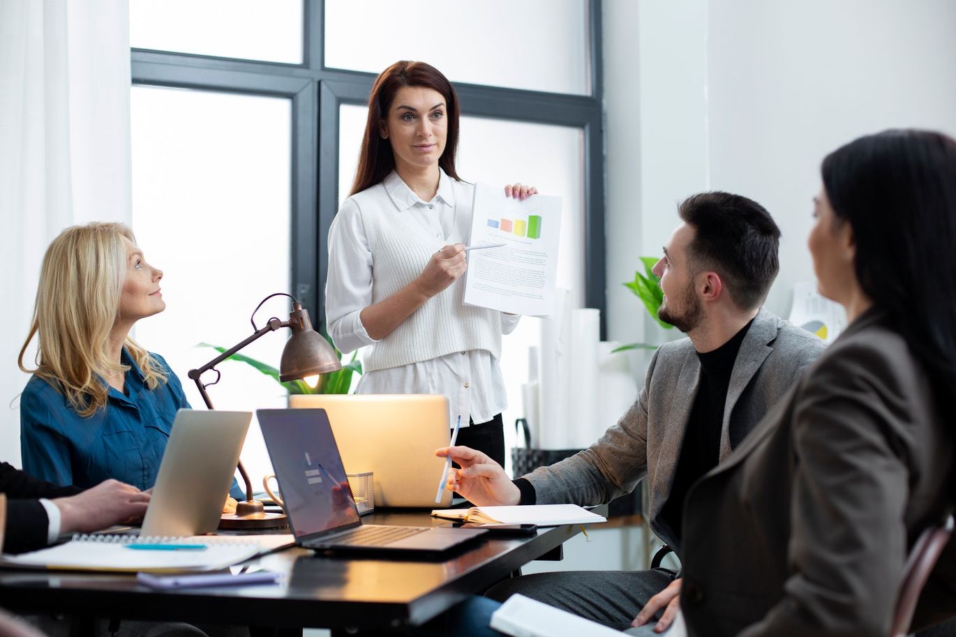 A group of people are sitting around a table with laptops and a woman is giving a presentation.