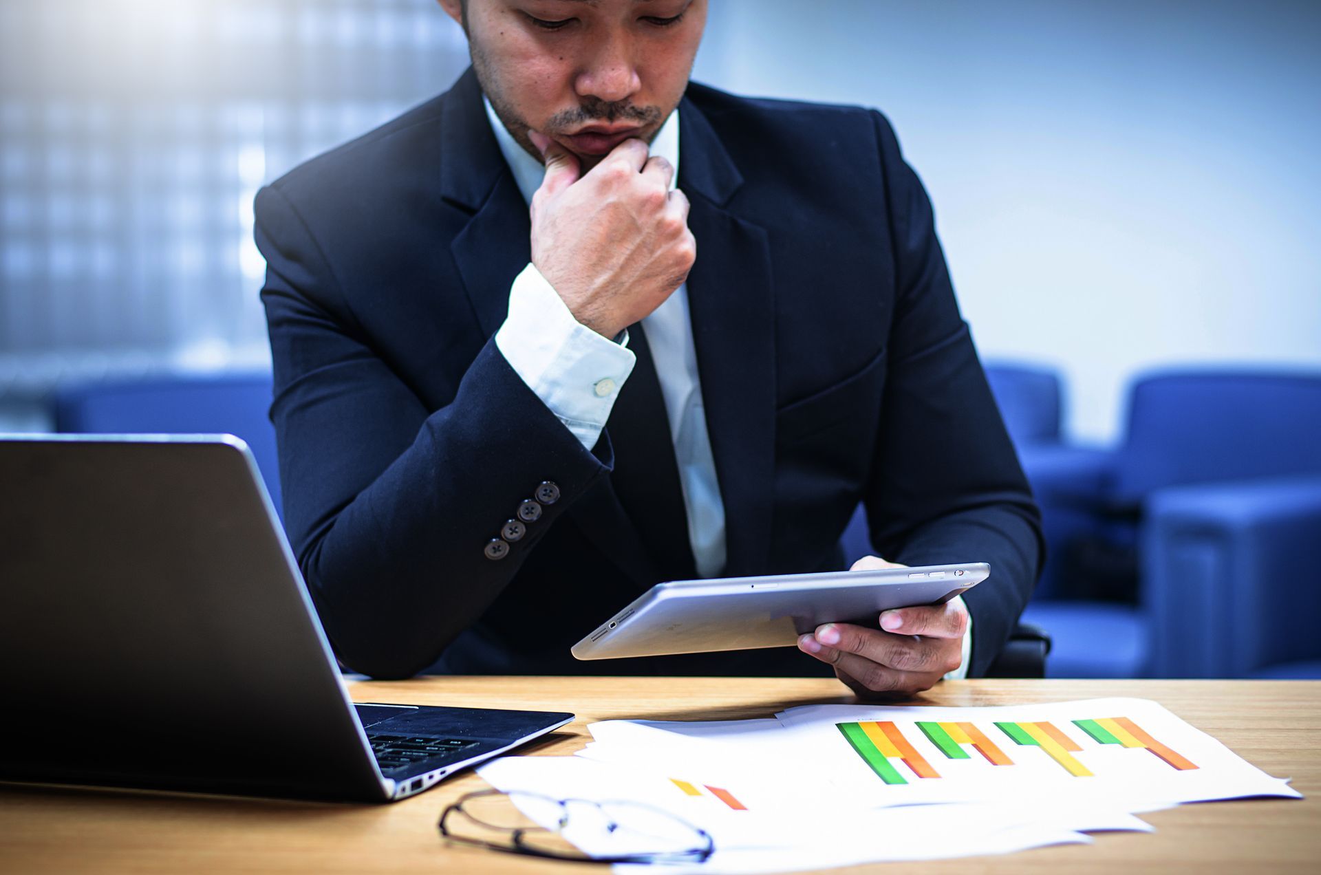 Man in suit examines tablet and charts, pondering at a desk with a laptop.
