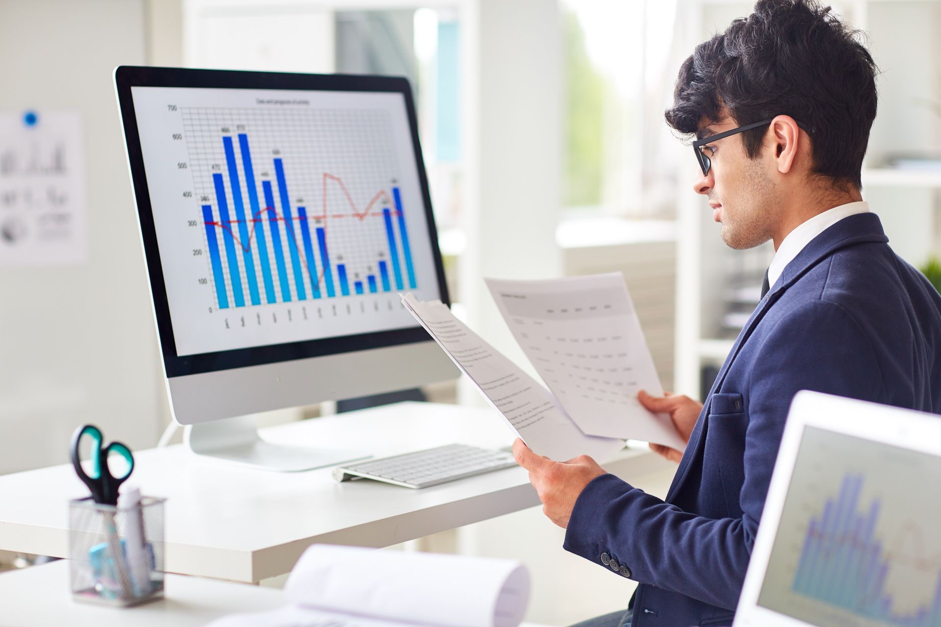 A man in a suit analyzes financial data on a computer screen and papers in an office setting.