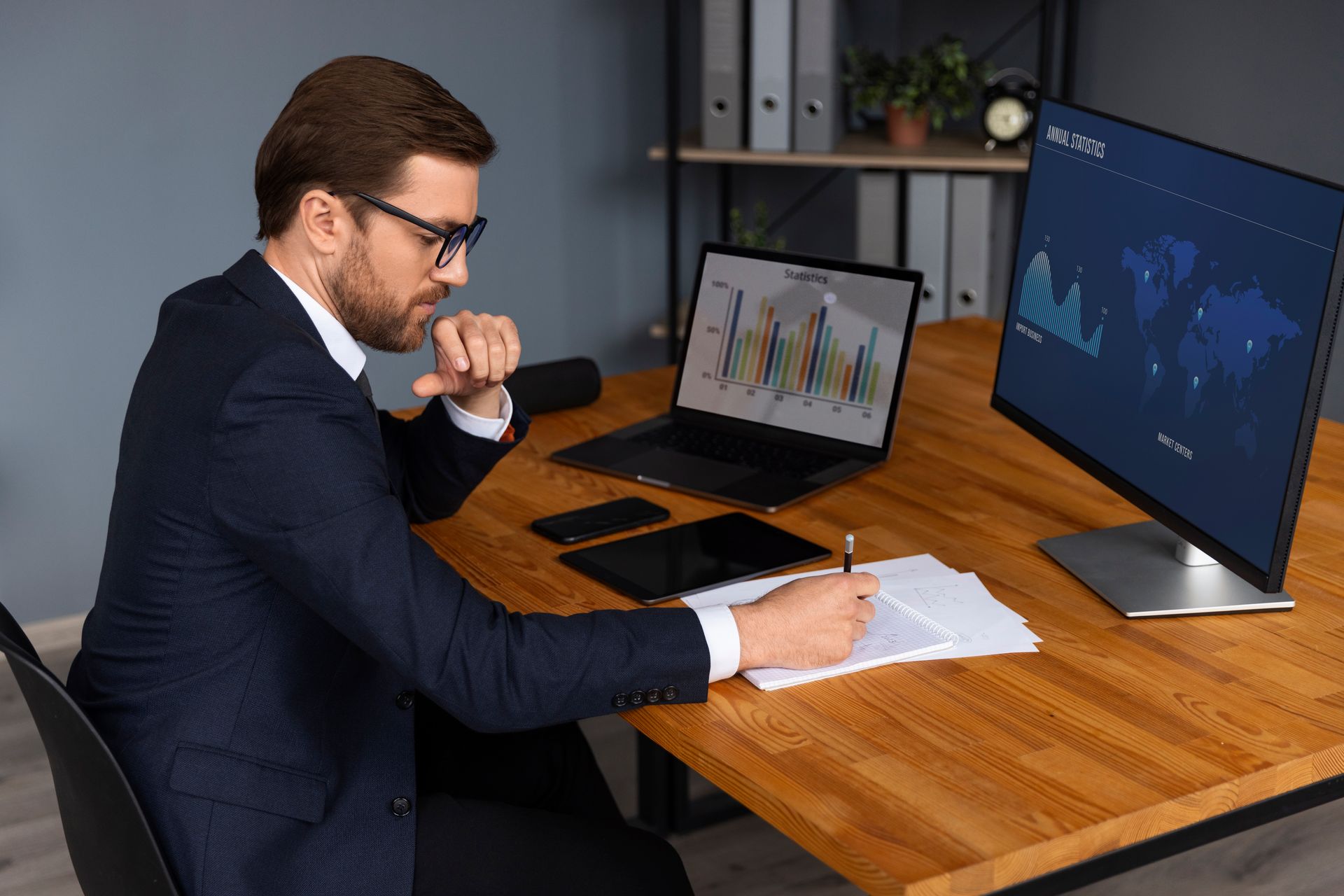 Man in suit at desk, analyzing data on laptop and monitor, writing on paper, office setting.