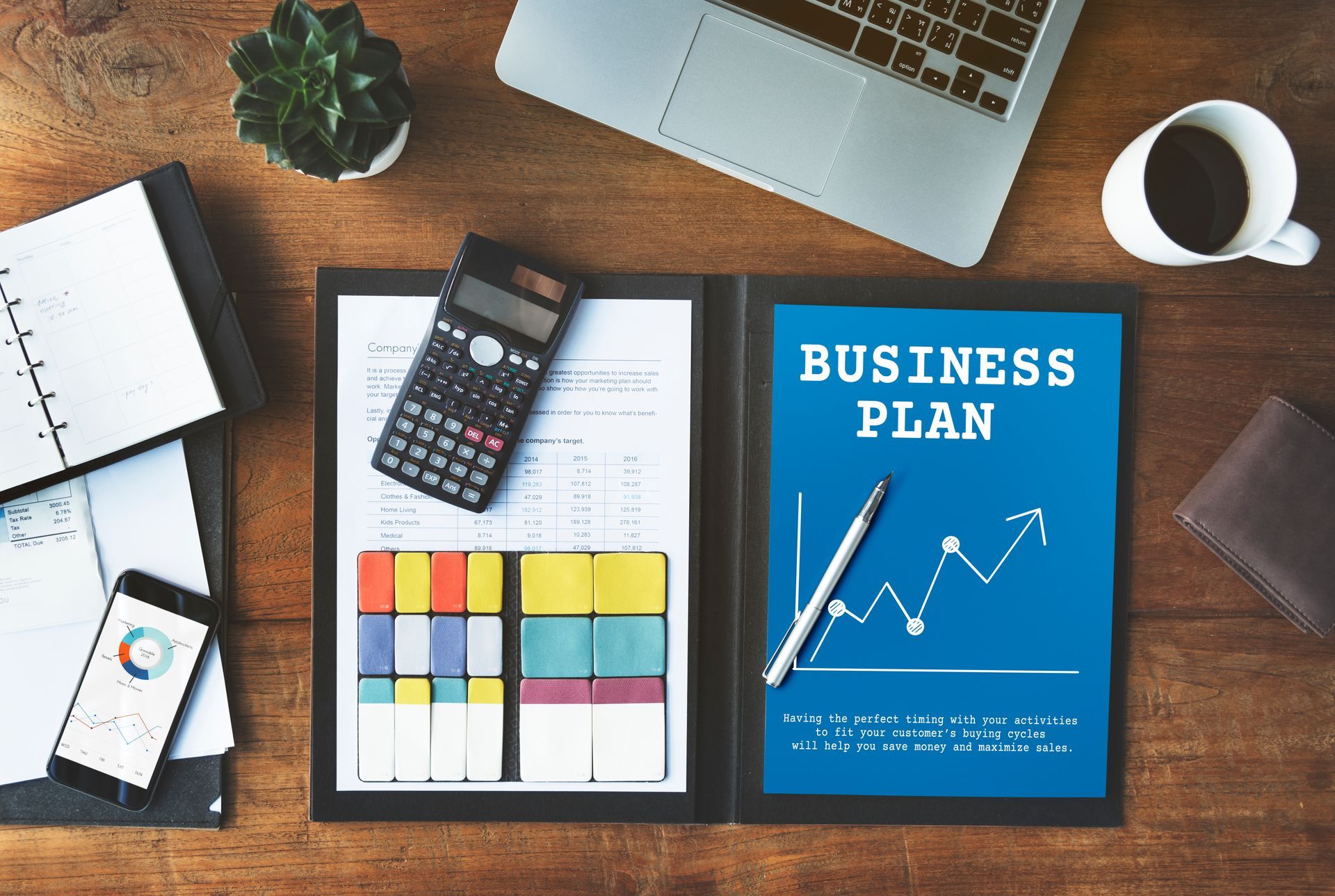 Business plan spread open on a wooden desk with calculator, phone, laptop, coffee, and a notebook.