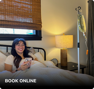 Person in bed with IV drip, holding phone, smiling. Interior setting with lamp, blinds, and nightstand.