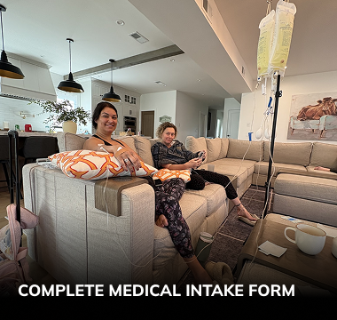 Two people receive IV infusions on a couch in a home setting. A bag hangs from a stand.
