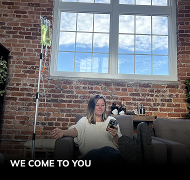 A person sits in an armchair, looking at their phone while receiving an IV drip in a room with a brick wall.