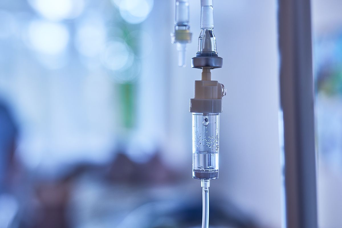 A close-up of a clear medical IV drip chamber attached to tubing, set against a blurred, clinical background.