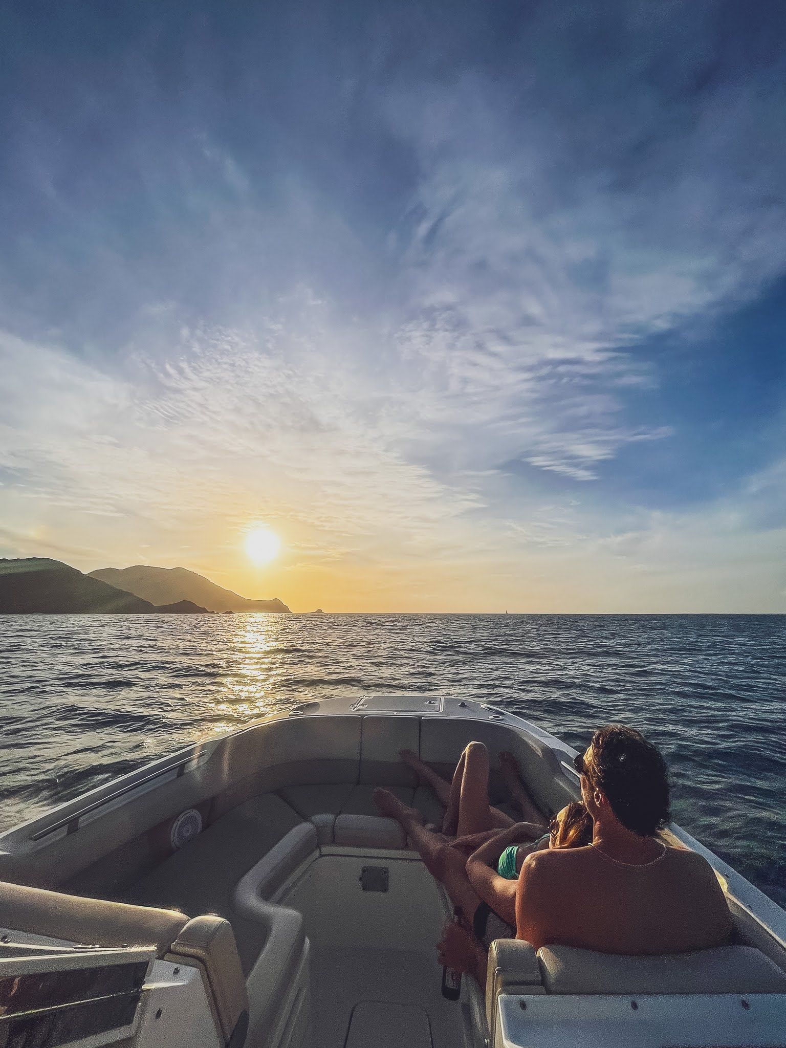 A man is sitting on the back of a boat in the ocean at sunset.