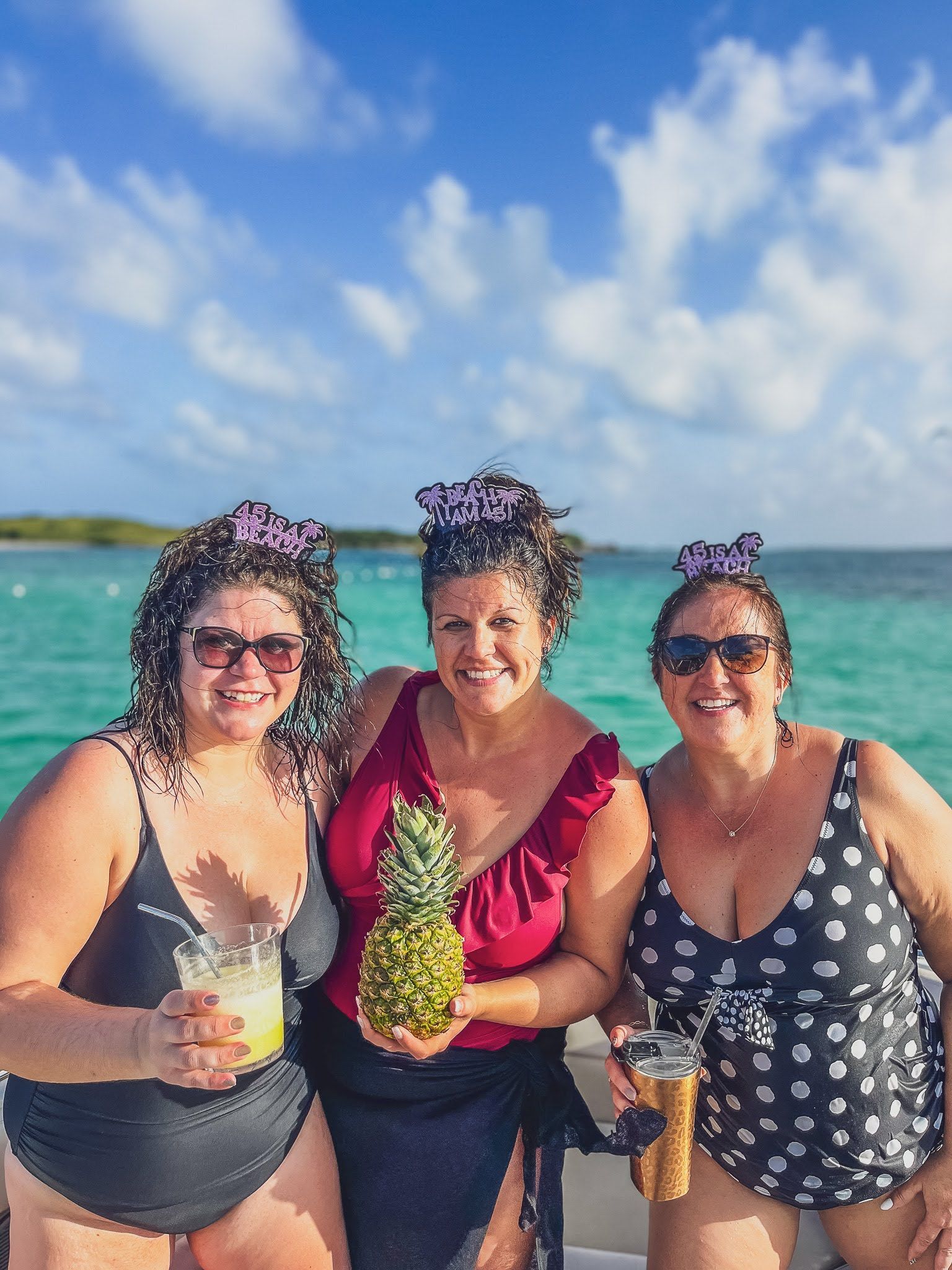 Three women in swimsuits are standing next to each other on a boat holding pineapples and drinks.