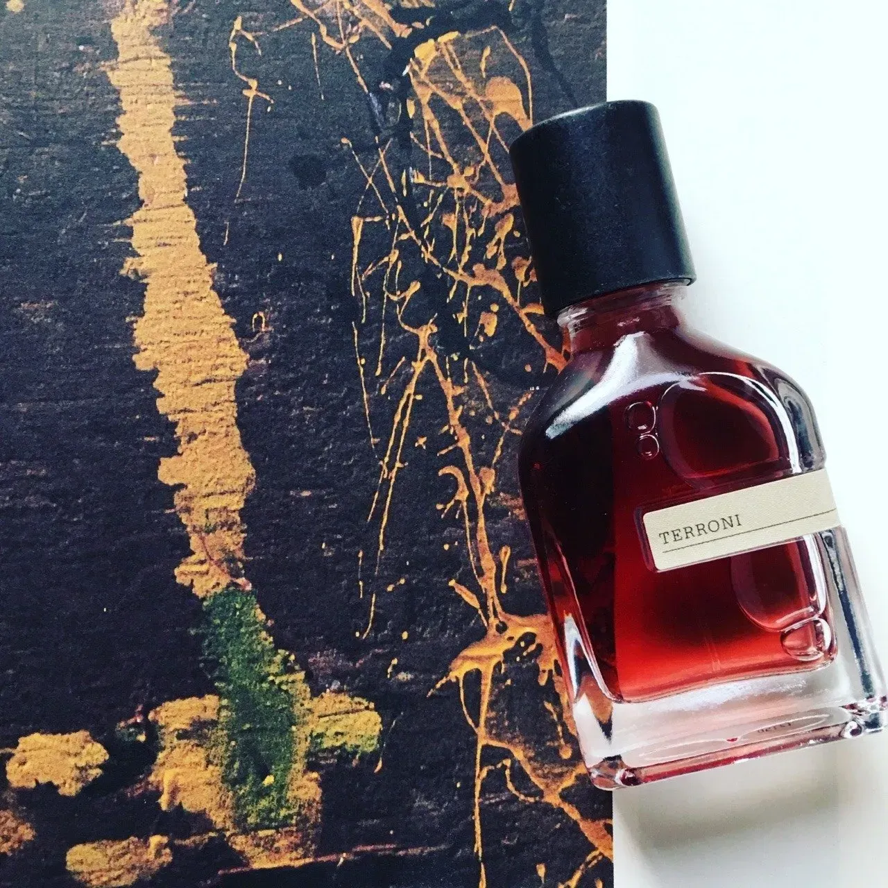 Flacone di profumo rosso con tappo nero su sfondo scuro e strutturato con striature di vernice dorata.