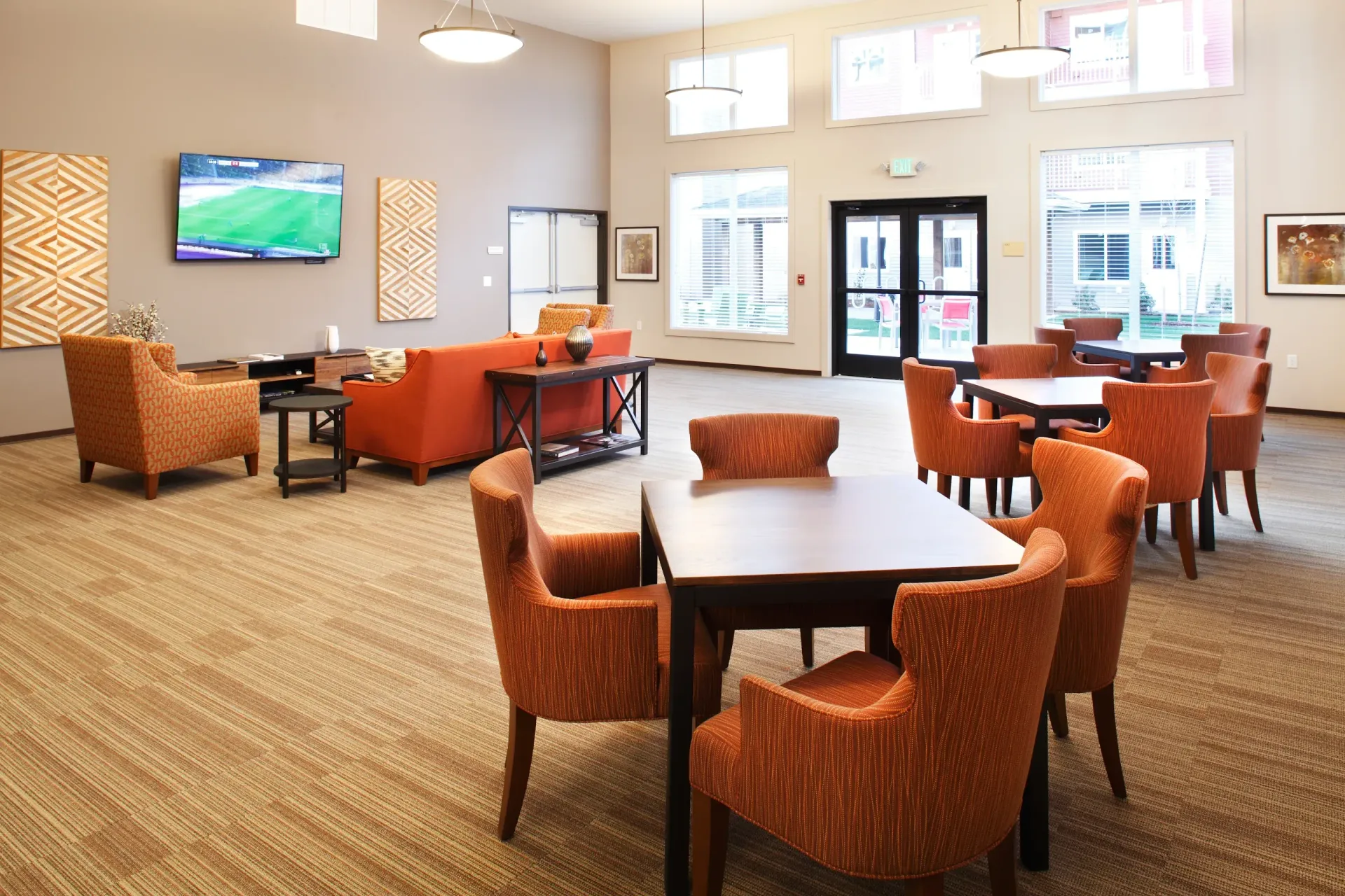 Spacious apartment community lounge with orange chairs, couches, tables, and a wall-mounted TV.