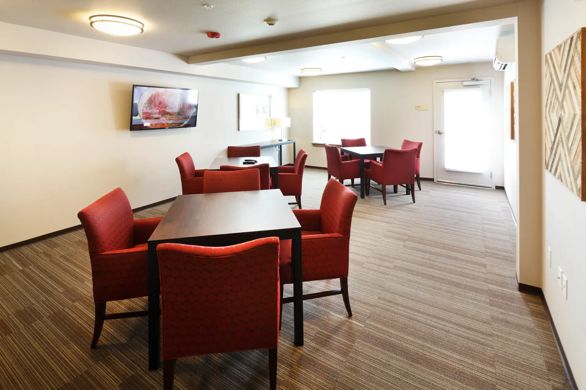 Common lounge area with red upholstered chairs, tables, a wall-mounted TV, and large windows.