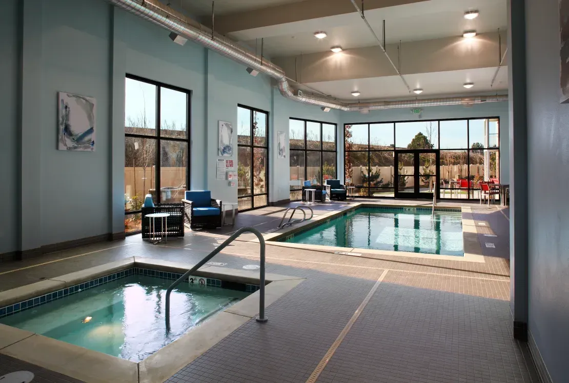 Indoor apartment community pool area with spa, seating, and large windows.