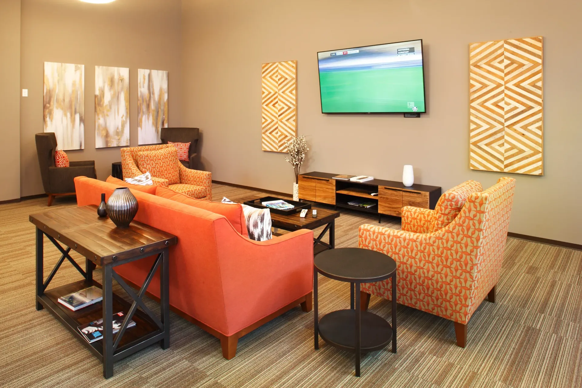 Modern community lounge with orange sofa, patterned chairs, coffee table, and wall-mounted TV.