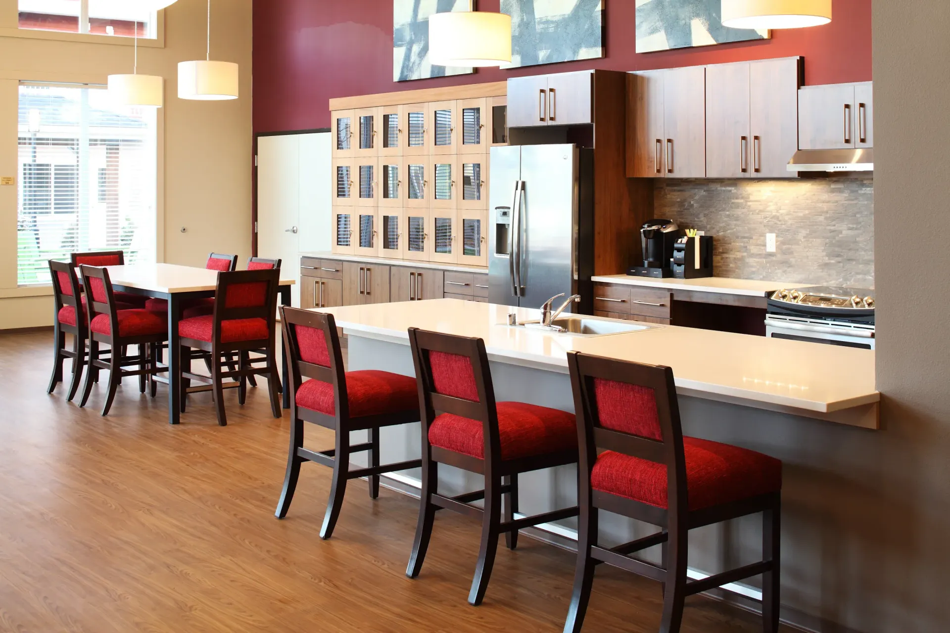 Modern communal kitchen with island, dark wood cabinets, red chairs, and stainless appliances.