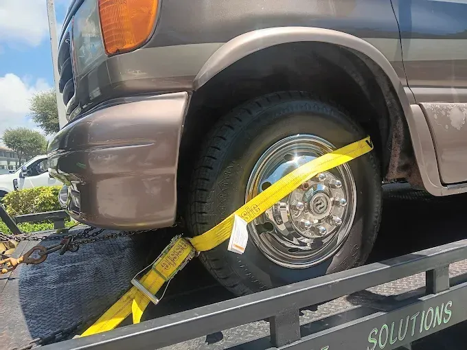 A tan van is secured to a flatbed tow truck with a yellow ratchet strap wrapped around its front tire.