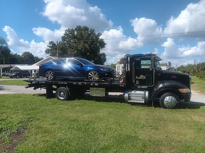 A blue sedan sits on the bed of a black flatbed tow truck parked on a grassy lot under a sunny sky.