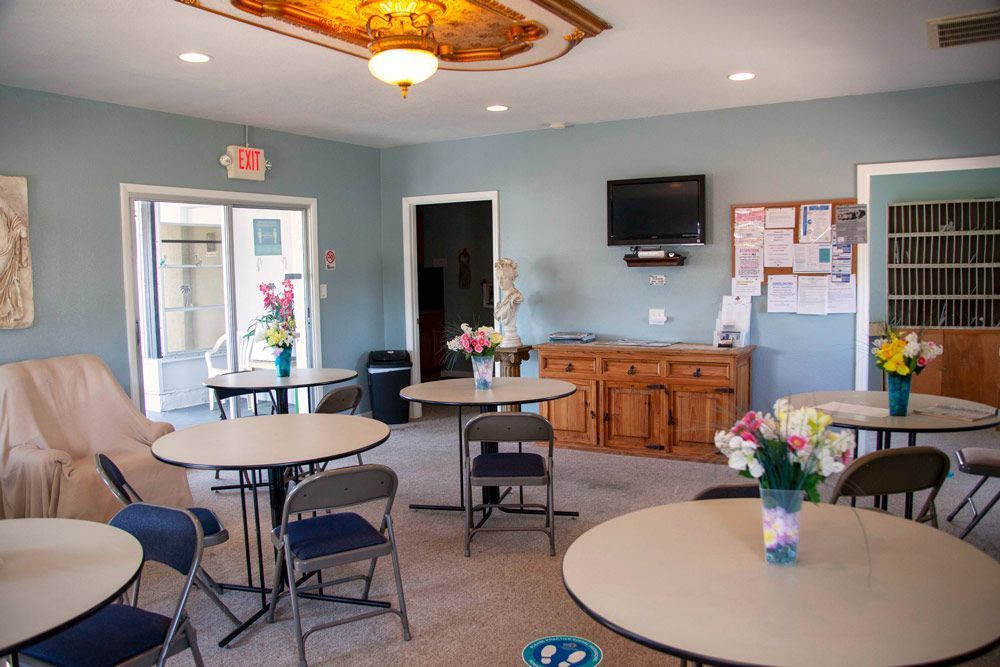 A lounge with round tables, chairs, flowers, TV, bulletin board, and exit door.