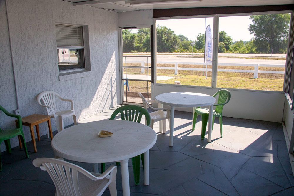 Enclosed porch with white plastic tables and chairs. View of road and green space through the screen.